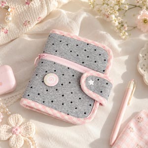 May include: A grey fabric notebook cover featuring a black star pattern, trimmed with pink and white gingham fabric. It includes a zipped pocket, a snap-button closure with a white star detail, and a circular pin with a pink swirl design.