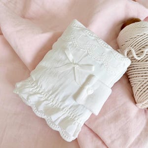 Könnte beinhalten: Ein weißes, rechteckiges Kissen mit Rüschenkante und Spitzendetails. Eine kleine weiße Schleife ist mittig auf der Vorderseite angebracht. Das Kissen liegt auf einer rosa Stoffoberfläche, mit einer Spule weißen Fadens im Hintergrund.