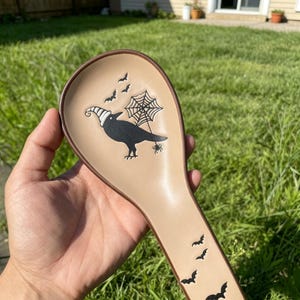 Könnte beinhalten: Beige Keramik-Löffelablage mit braunem Rand, mit einer schwarzen Krähe mit Hexenhut, einem Spinnennetz und Fledermäusen. Die Löffelablage ist für Halloween und den Küchengebrauch konzipiert.