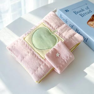 May include: A quilted pink fabric pouch featuring a light green apple appliqu&eacute; on a square yellow patch. The pouch includes a small attached flap with a white star detail and a zip closure, positioned next to a blue book titled Book of Bread.