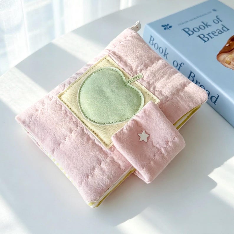 May include: A quilted pink fabric pouch featuring a light green apple appliqu&eacute; on a square yellow patch. The pouch includes a small attached flap with a white star detail and a zip closure, positioned next to a blue book titled Book of Bread.
