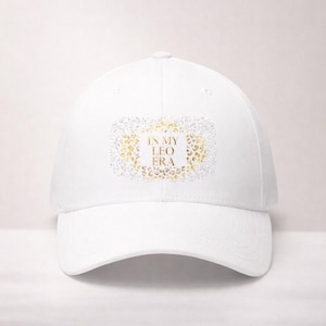 Könnte beinhalten: Weiße Baseballkappe mit gebogenem Schirm. Auf der Kappe steht der Text "IN MY LEO ERA" in goldfarbenen Buchstaben, umgeben von einem goldenen und silbernen Glitzerdesign. Ein klassisches, lässiges Accessoire.