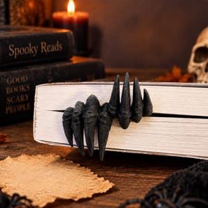 May include: A black bookmark shaped like a clawed hand with long, pointed fingers. The bookmark is inserted into the pages of a closed book, with the fingers extending over the edge of the pages. The texture of the hand is matte and dark.