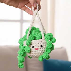 May include: A crocheted hanging planter with a smiling face, filled with green, curly, plant-like yarn. The planter is off-white with black eyes and a red mouth. The hanging cord is also off-white.