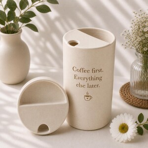 以下が含まれることがあります： ベージュ色のトラベルマグと、それに合う蓋。マグには「Coffee first. Everything else later.」の文字とコーヒーカップのグラフィックが描かれています。マグは花の入った花瓶の隣にあります。