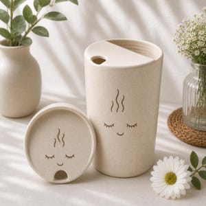 May include: A beige travel mug with a matching lid. The mug and lid feature a whimsical design of closed eyes and steam rising from the top. The mug is made of a textured material and is suitable for hot or cold drinks.