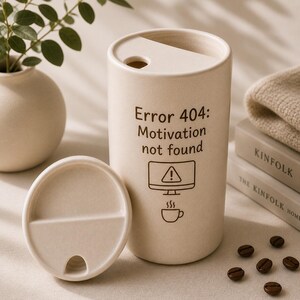 May include: A beige travel mug with a lid, featuring the text "Error 404: Motivation not found" and a computer screen icon. The mug is next to its lid, a small vase with greenery, and a stack of books.
