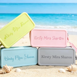Peut inclure: Quatre pochettes rectangulaires en silicone avec dragonnes, déclinées en vert citron, rose pastel, bleu clair et gris. Chaque pochette porte le texte personnalisé Kirsty Mira Sharon imprimé sur le devant dans une police assortie.