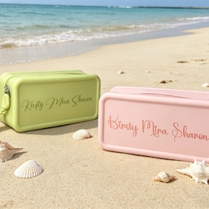 Peut inclure: Une pochette rectangulaire en silicone vert citron et une pochette rectangulaire en silicone rose clair, chacune dotée d'une fermeture éclair, d'une dragonne et du texte personnalisé Kirsty Mina Sharon imprimé en foncé sur le devant.
