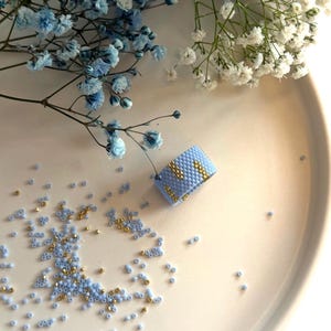 Op de afbeelding: Een brede ring gemaakt van lichtblauwe kraaltjes in een geweven patroon, met verticale accenten van metallic gouden kraaltjes. De ring is omringd door losse lichtblauwe en gouden kraaltjes, met takjes blauwe en witte gipskruidbloemen erbij.