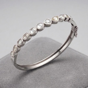 Può includere: Un bracciale rigido in argento con una fila di pietre preziose chiare e sfaccettate. Il bracciale ha una chiusura a cerniera ed è appoggiato su una superficie grigia. Le pietre preziose hanno forme diverse, aggiungendo un tocco unico al gioiello.