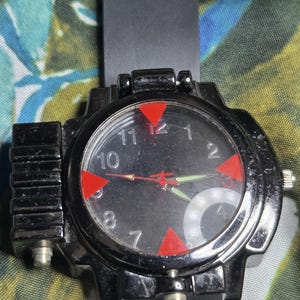 May include: A black wristwatch with a round face, featuring a black dial with white numerals, red triangular markers at the 12, 3, 6, and 9 o'clock positions, and red and green hands. The watch has a black silicone strap and a black plastic case.