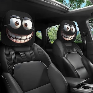 May include: Black car headrest covers with a cartoon face design. The headrests feature a smiling face with large white teeth, big eyes, and a black background. The car seats are black leather.