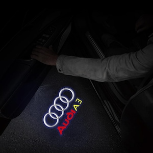 May include: A car door projects a glowing Audi A3 logo onto the ground. The logo shows four interlocking rings in white, with "Audi A3" in red. The image is taken at night, with a person's hand visible near the open door.