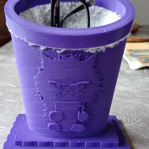 May include: A purple 3D-printed glasses holder with a cat design. The holder is cylindrical with a square base and holds black-framed glasses. The cat's face is embossed on the front. The holder is approximately 10 cm tall.