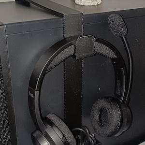 May include: Black over-the-ear headphones with a built-in microphone are suspended from a black metal headphone stand. The stand is mounted on a black computer tower. The headphones have the brand name "Trust" on them.