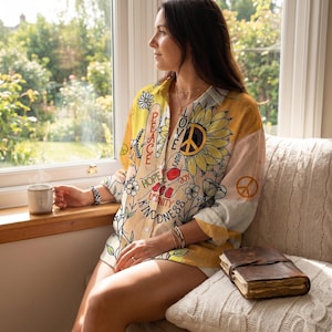 Puede incluir: Camisa estampada con girasoles y símbolos de la paz. La camisa incluye las palabras «Paz», «Amor», «Esperanza», «Unidad» y «Bondad». La camisa es de color amarillo, blanco y melocotón.