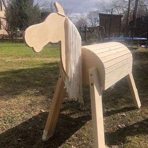 Peut inclure: Un cheval à bascule en bois avec une finition marron clair. Le cheval présente un corps incurvé, quatre pattes et une tête avec une crinière blanche flottante. Le cheval est conçu pour le jeu en extérieur et est posé sur une pelouse.