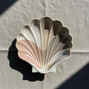 Puede incluir: Un plato de cerámica con forma de concha con borde festoneado. El plato presenta una paleta de colores blanco, rosa, beige y marrón, con un acabado brillante. El plato está diseñado para parecerse a una concha y probablemente se utiliza con fines decorativos.