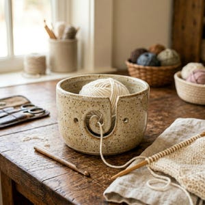 May include: A speckled beige ceramic yarn bowl with a spiral cutout on the front, containing a ball of cream-colored yarn. The bowl features a smooth, rounded shape with a flat base, designed to hold yarn while knitting or crocheting.