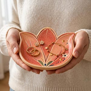 May include: A lotus-shaped ceramic trinket dish in shades of coral and pink with speckled details. The dish features painted flower and star motifs and holds two gold-toned rings, a delicate gold chain bracelet, and two small flower-shaped studs.