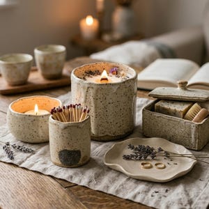 May include: A collection of speckled ceramic items including a large candle with a wooden wick, a small candle, a matchstick holder with matches, a lidded rectangular box containing a soap bar and brush, and a small scalloped dish with two gold rings.