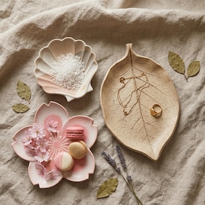 May include: A collection of three ceramic dishes: a shell-shaped bowl containing white salt, a pink cherry blossom-shaped plate holding three macarons and small flowers, and a large speckled leaf-shaped tray holding a gold necklace and ring.