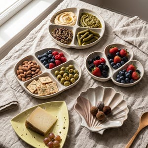 May include: A collection of ceramic serving dishes including two round four-section plates filled with hummus, pesto, ground meat, pickles, almonds, crackers, olives, and berries, a shell-shaped bowl with chocolates, and a triangular yellow plate.