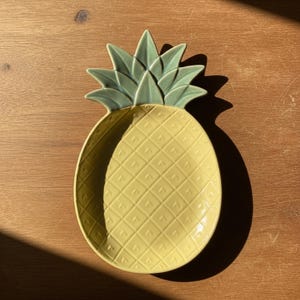 May include: A yellow ceramic pineapple-shaped serving dish with a textured surface and green leaves. The dish is designed with a diamond pattern and is ideal for serving snacks or as a decorative piece. The dish is on a wooden surface.