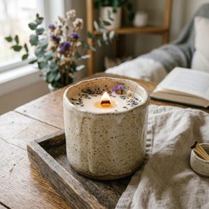 Pode incluir: Uma vela de cerâmica cor de creme com acabamento salpicado e efeito de esmalte escorrido. A vela apresenta um pavio de madeira aceso e é decorada com botões de lavanda secos e pequenas flores roxas, sobre uma bandeja de madeira.