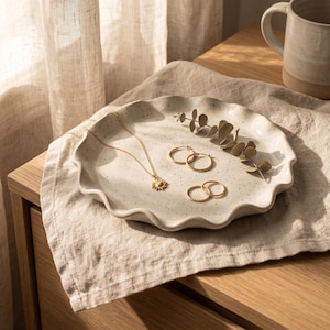 May include: A gold necklace with a sun pendant, two pairs of gold earrings, and a decorative tray with a wavy edge. The tray is speckled and holds a sprig of greenery. A beige mug is in the background.