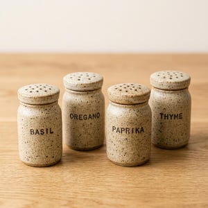 以下が含まれることがあります： 斑点のあるベージュの釉薬と穴の開いた蓋が付いた、4つの円筒形のセラミック製スパイスジャー。各ジャーには、Basil、Oregano、Paprika、Thymeと黒い文字で記されており、平らな面に一列に並べられています。
