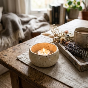 Puede incluir: Un cuenco de cerámica de color crema, de forma redonda y con motas de color marrón oscuro, que contiene una vela de té encendida. El cuenco tiene un borde grueso y liso, con una forma ligeramente irregular, sobre una superficie de tela.