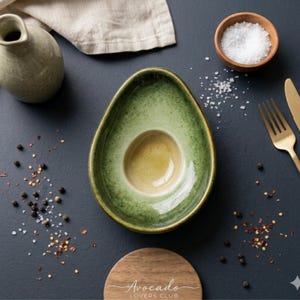 Puede incluir: Plato de cerámica con forma de aguacate, verde y amarillo. La imagen incluye un pequeño cuenco de madera con sal, pimienta, un tenedor y un cuchillo dorados, y un posavasos con el texto "Avocado Lovers Club".