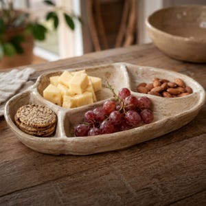 Op de afbeelding: Een beige, driedelig serveerplateau van keramiek met een onregelmatige, organische vorm. Het plateau bevat een stapel ronde crackers, blokjes gele kaas, een tros rode druiven en een klein hoopje bruine amandelen in aparte vakjes.