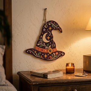 May include: A dark purple wall hanging shaped like a pointed hat, featuring a crescent moon, stars, a spider, and floral patterns in orange, green, and white. The hat has a tan band with a diamond-shaped buckle and hangs from a jute cord.