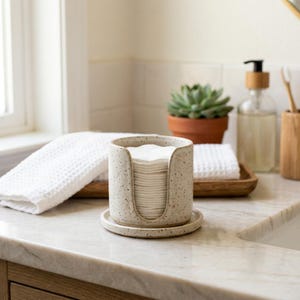Könnte beinhalten: Ein gesprenkelter Keramikhalter mit U-förmigem Ausschnitt, der einen Stapel runder weißer Wattepads enthält. Der Halter steht auf einer passenden runden Keramikuntertasse und weist eine hellbeige Basis mit dunkelbraunen Sprenkeln auf.