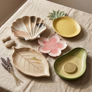 May include: A collection of five ceramic dishes shaped like a seashell with tan and brown accents, a yellow pineapple, a pink cherry blossom, a speckled beige leaf, and a green avocado, accompanied by a small bundle of dried lavender sprigs.