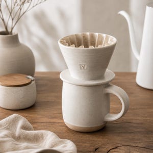 May include: Cream-colored ceramic coffee pour-over set with a matching mug. The set includes a cone-shaped dripper with a paper filter, a mug with a handle, and a small, round container with a wooden lid. The mug has a light brown base.