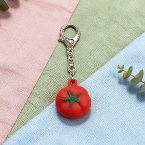 May include: A red tomato-shaped keychain with a green star-shaped design on top. The keychain has a silver-colored clasp and chain. The background is a mix of pink, green, and blue fabric.