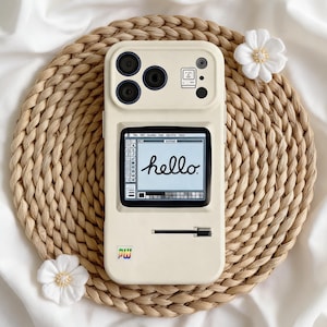 May include: A cream-colored phone case with a retro computer design. The case features a screen displaying the word "hello" in cursive, along with a small logo and camera lenses. The case is on a woven mat with white flowers.