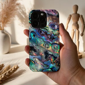 May include: A smartphone case with a vibrant, iridescent abalone shell design. The case features a multi-lens camera cutout and a glossy finish, showcasing a blend of blues, greens, and purples. The case is held in a hand, with a white background.