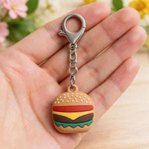 May include: A colorful burger-shaped keychain with a silver clasp and chain. The burger has a sesame seed bun, red tomato, yellow cheese, brown patty, and green lettuce. The keychain is held in a hand.