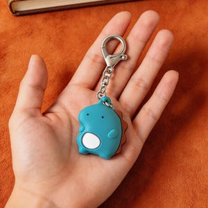 May include: A teal dinosaur keychain with a silver clasp and chain. The dinosaur has a white belly and brown accents. The keychain is held in a hand against an orange background, with a brown book in the background.