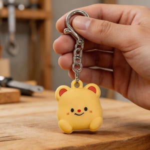 May include: A yellow bear-shaped keychain with a silver clasp and chain. The bear has red accents on its ears and a smiling face with a small red nose. The keychain is held against a wooden surface.