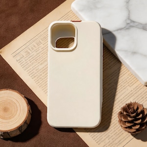 May include: A cream-colored phone case with a camera cutout, resting on a vintage newspaper. A small wooden slice, a marble square, and a pinecone are also present. The case is smooth and appears to be made of a soft material.