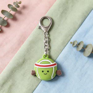 May include: A green, cartoon-style keychain featuring a smiling character with a red headband and brown arms. The keychain has a silver clasp and chain, and is set against a backdrop of pink, green, and blue fabric.