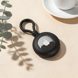 以下が含まれることがあります： キーやバッグに取り付けるためのループが付いた、黒い丸いAirTagホルダー。ホルダーは、Appleのロゴが入った透明な窓を備えています。背景にはコーヒーカップと緑の小枝があります。
