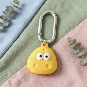 May include: A yellow, cartoon-faced keychain with a silver carabiner clip. The keychain has a rounded shape with two large, white-eyed circles and small dimples. The background is a mix of blue, green, and pink fabric.