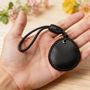 May include: A black leather key fob with a stitched border and a black cord loop. The fob is round with a small metal ring at the top. The background is blurred, with hints of flowers and wood.
